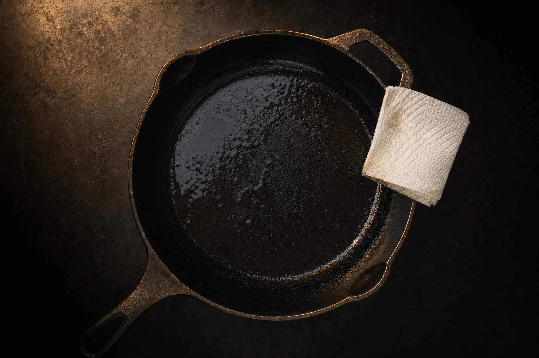 Cast iron skillet with an uneven sticky sheen of half-cured oil, paper towel resting on the rim